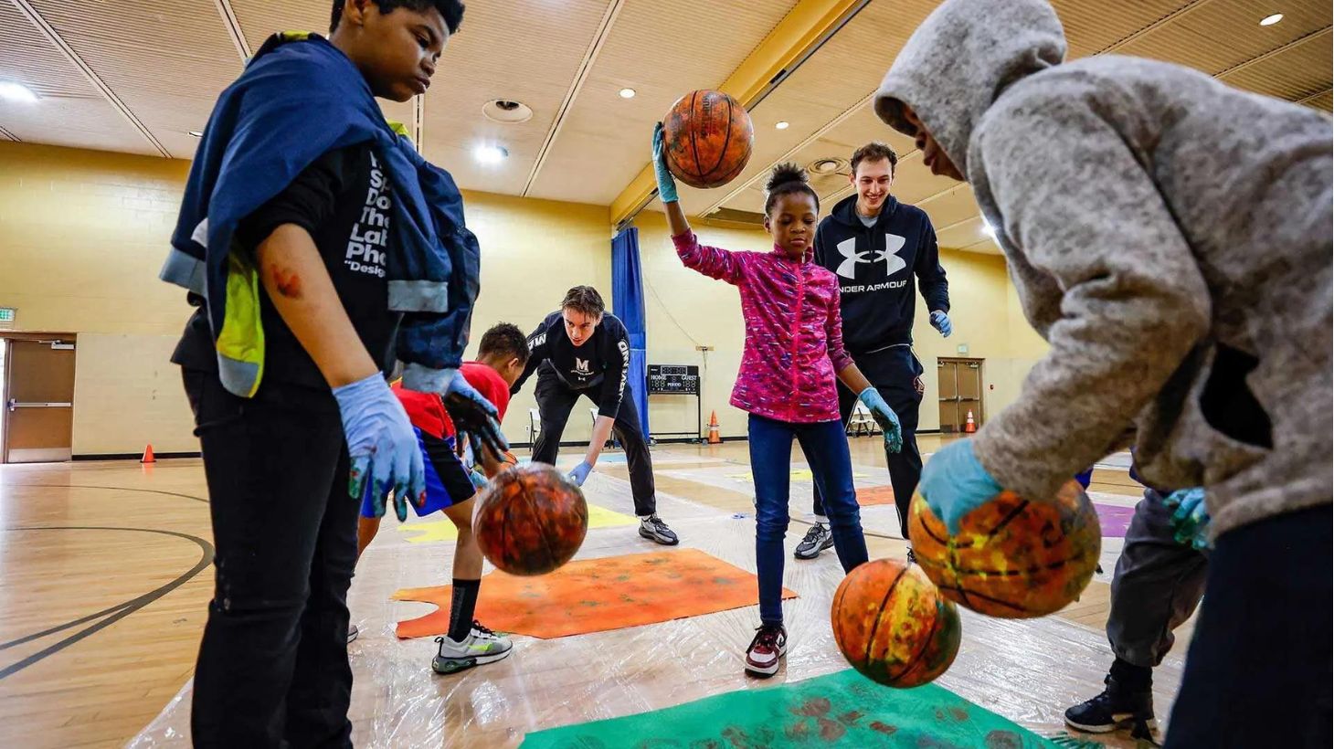 UMD basketball players and art professor help children to paint with paint-smeared basketballs in a gym.