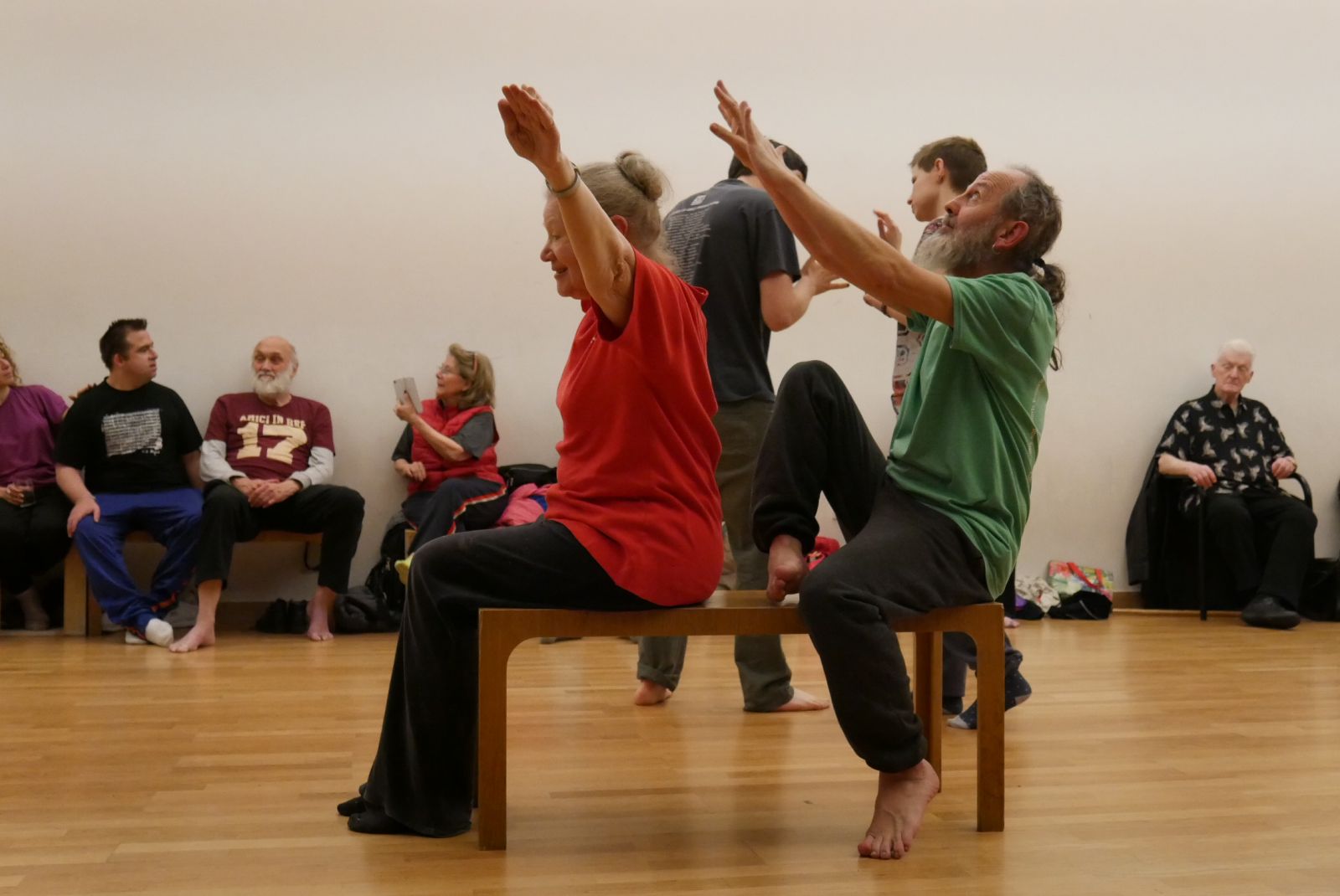 Two dancers at London’s Amici Dance Theatre Company sit on a bench. One woman reaches her arms to the sky and behind her a man spirals to reach toward her, head tipped up. Around them people sit and watch, and behind them two more dancers are touching hands, gently. 