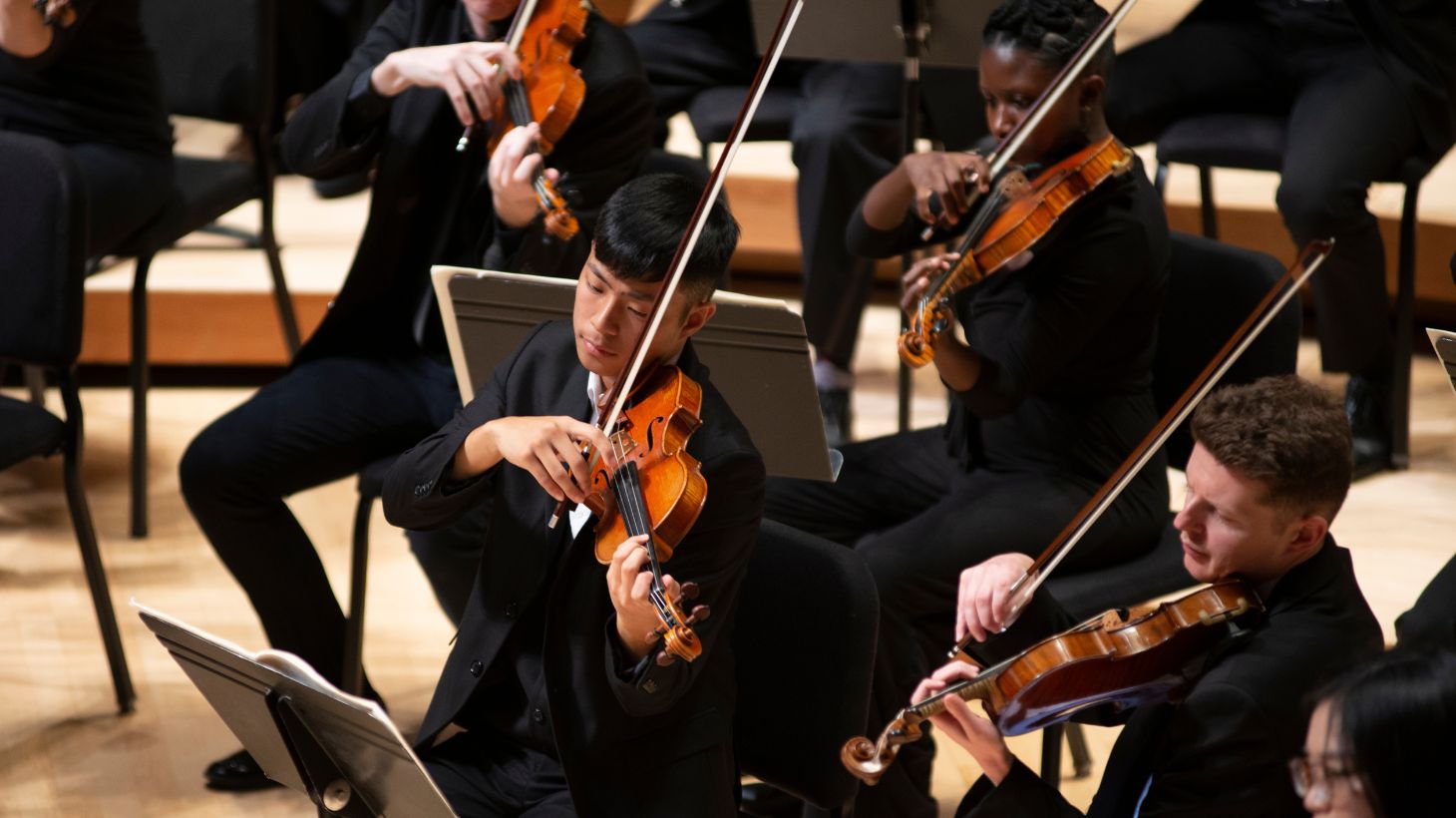 Men and women in an orchestra play violins on stage.