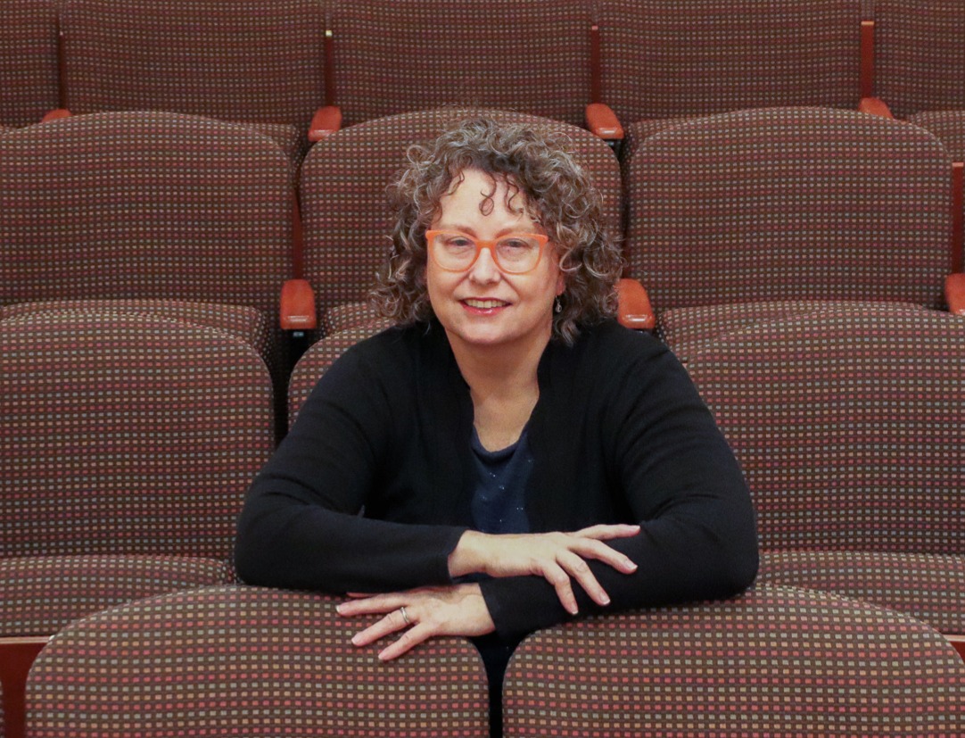 Picture of a white women with short curly brown hair seated in an auditorium. 