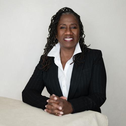 Sherrilyn Ifill, a confident Black woman dressed in a white dress shirt and black suit jacket with her hands folded on a table