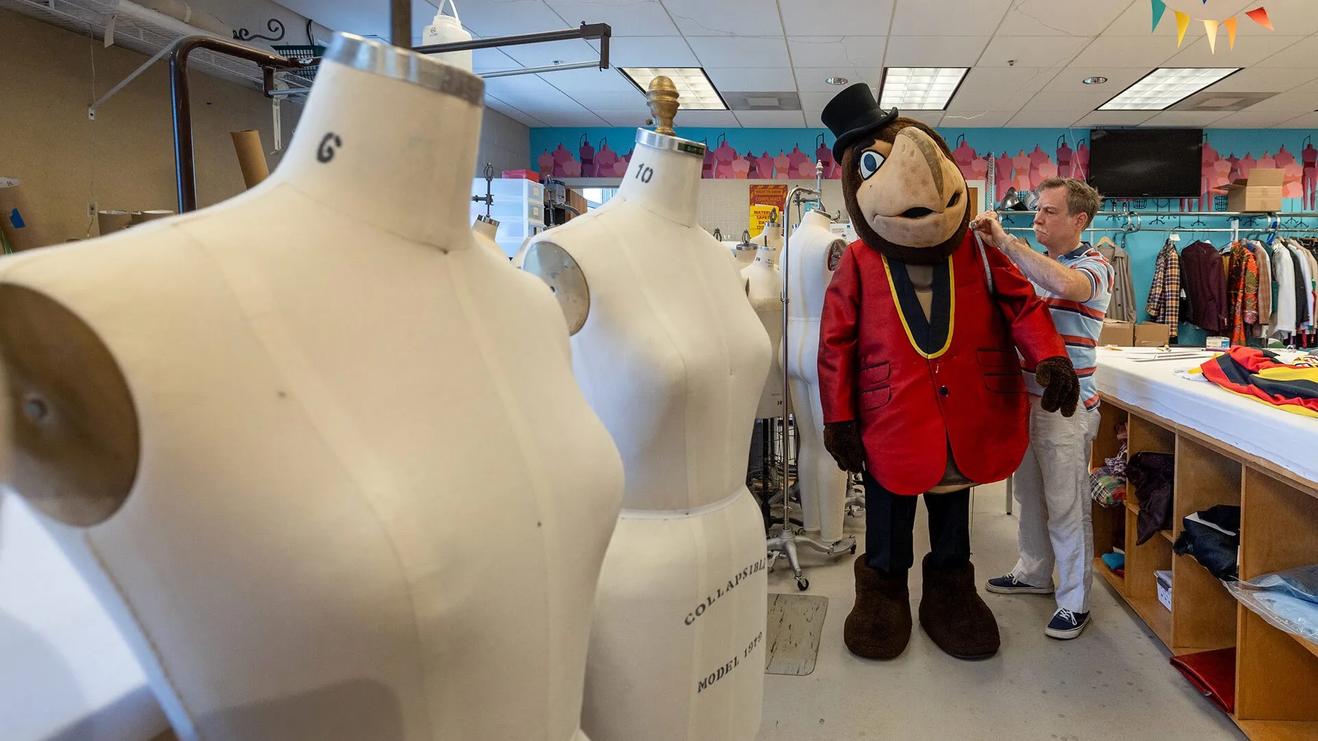 Costume manequins are featured in the foreground. In the back, a man is measuring Testudo's new jacket.