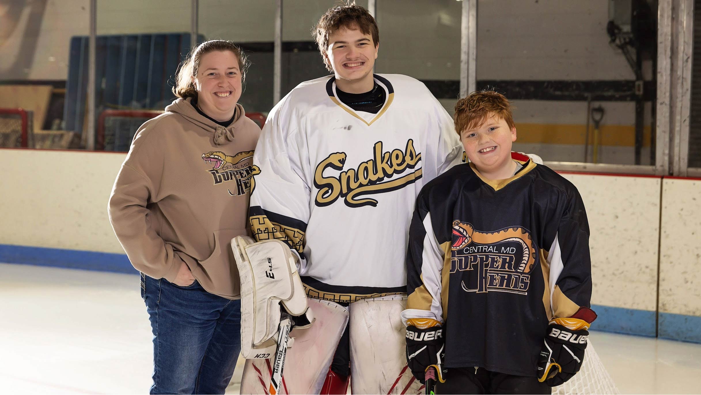 A smiling adult stands on an ice rink with two young hockey players. The adult wears a tan hoodie with a snake-themed team logo. The taller player in the middle is wearing white goalie gear and a white jersey that says “Snakes,” while the younger player on the right wears a black jersey with a similar snake logo. All three are posing together in front of the boards on the rink.