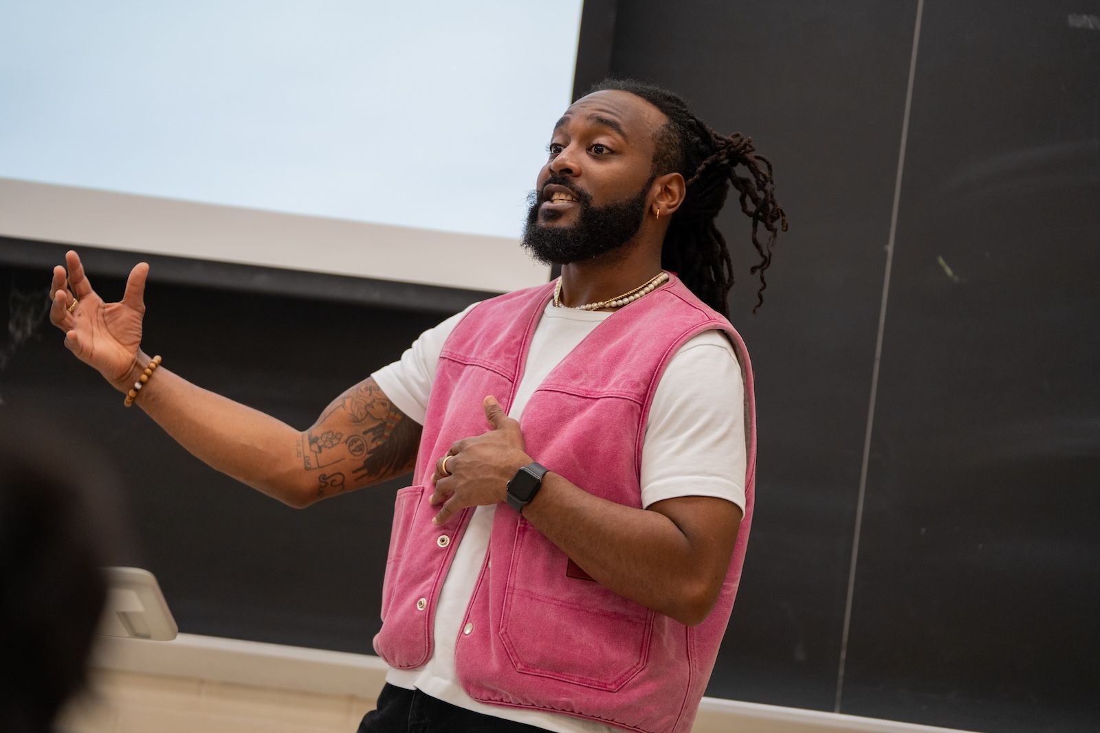 A man stands teaching in front of a chalkboard and screen. He is wearing a white t-shirt and pink vest.