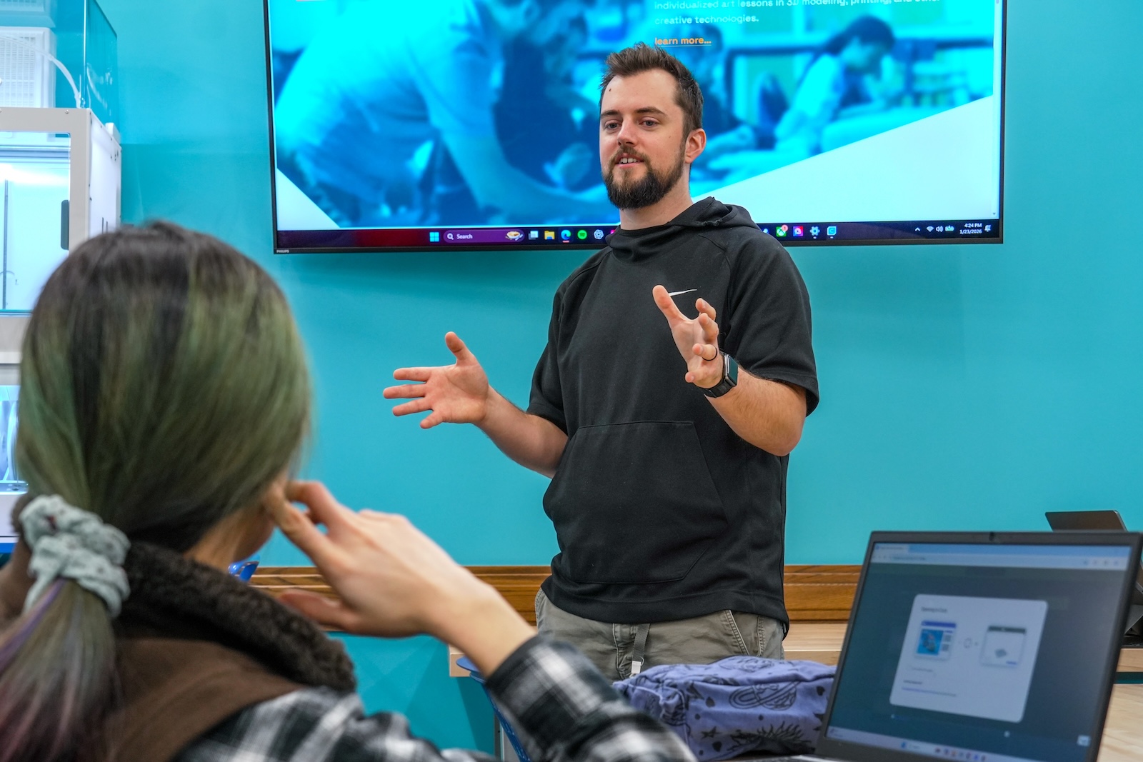 A man is speaking in front of a screen to a woman who is looking on.
