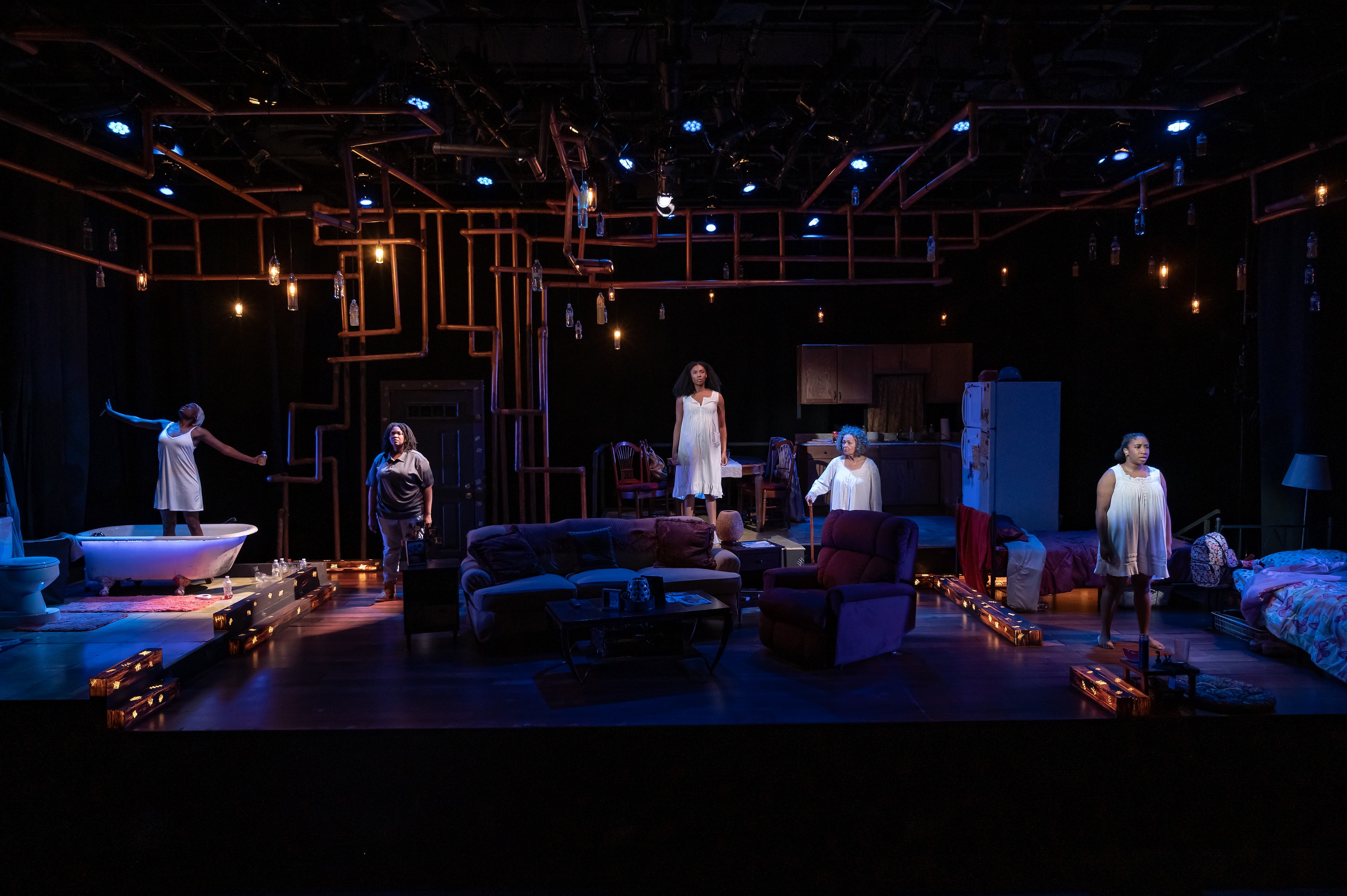 A theatrical stage set depicting a domestic scene with five actors in various poses. The stage is dimly lit with overhead blue and amber lights. On the left, an actor stands near a bathtub, gesturing outward. In the center, two actors stand near a door and a kitchen area, respectively. On the right, two more actors are positioned near a sofa and a bed. The set includes furniture like sofas, a table, and a bed, creating a home-like atmosphere. Exposed piping and hanging lights add to the visual complexity of