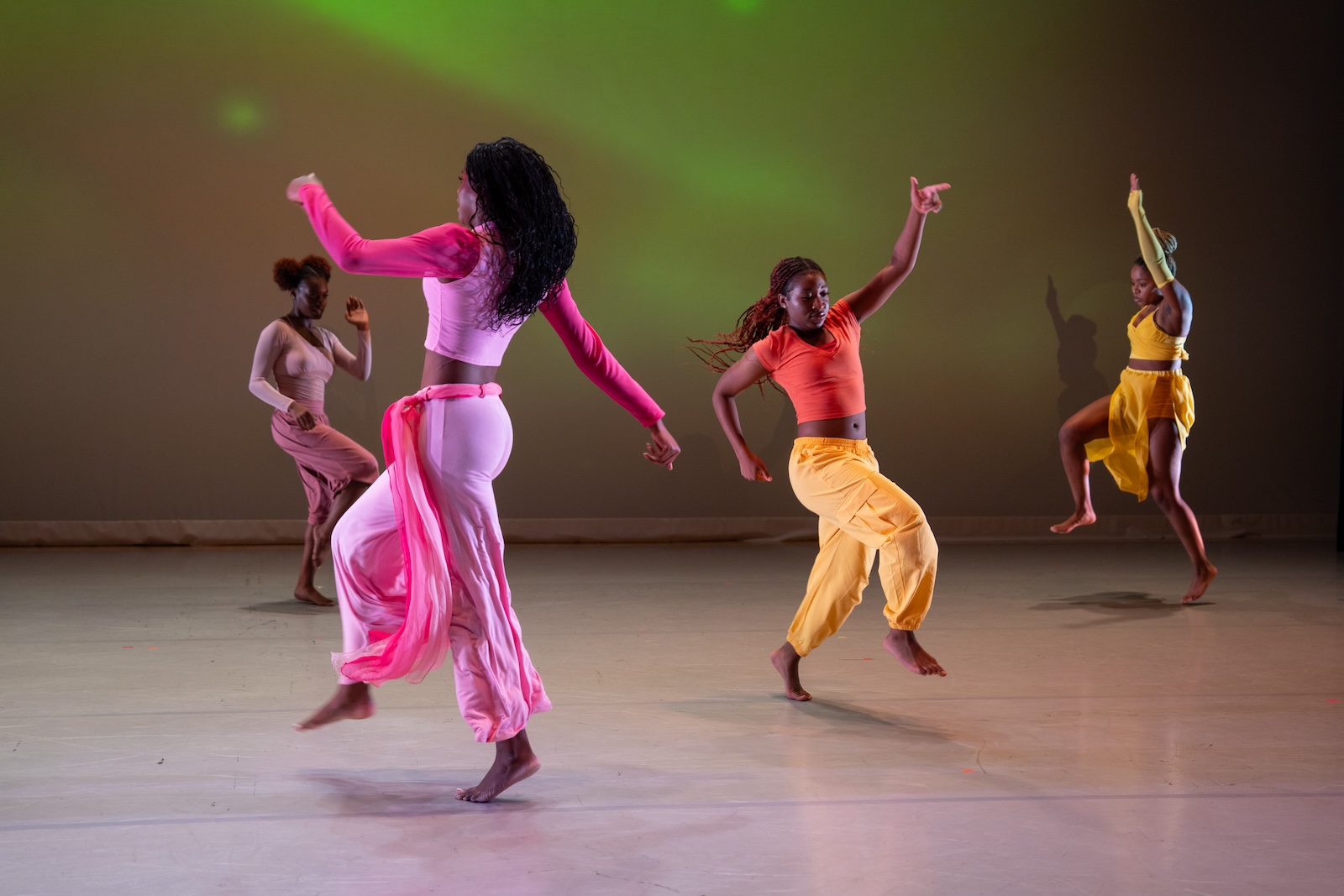 Four women dressed in vibrant colors hop and dance across a stage with their arms waving in the air during a performance. 