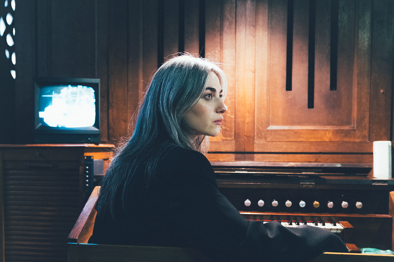 Kali Malone sits at an organ. There is a soft white light shining from above, illuminating part of the organ and the side profile of Malone's face. There is an old tv sitting behind her, with an image displayed on it.