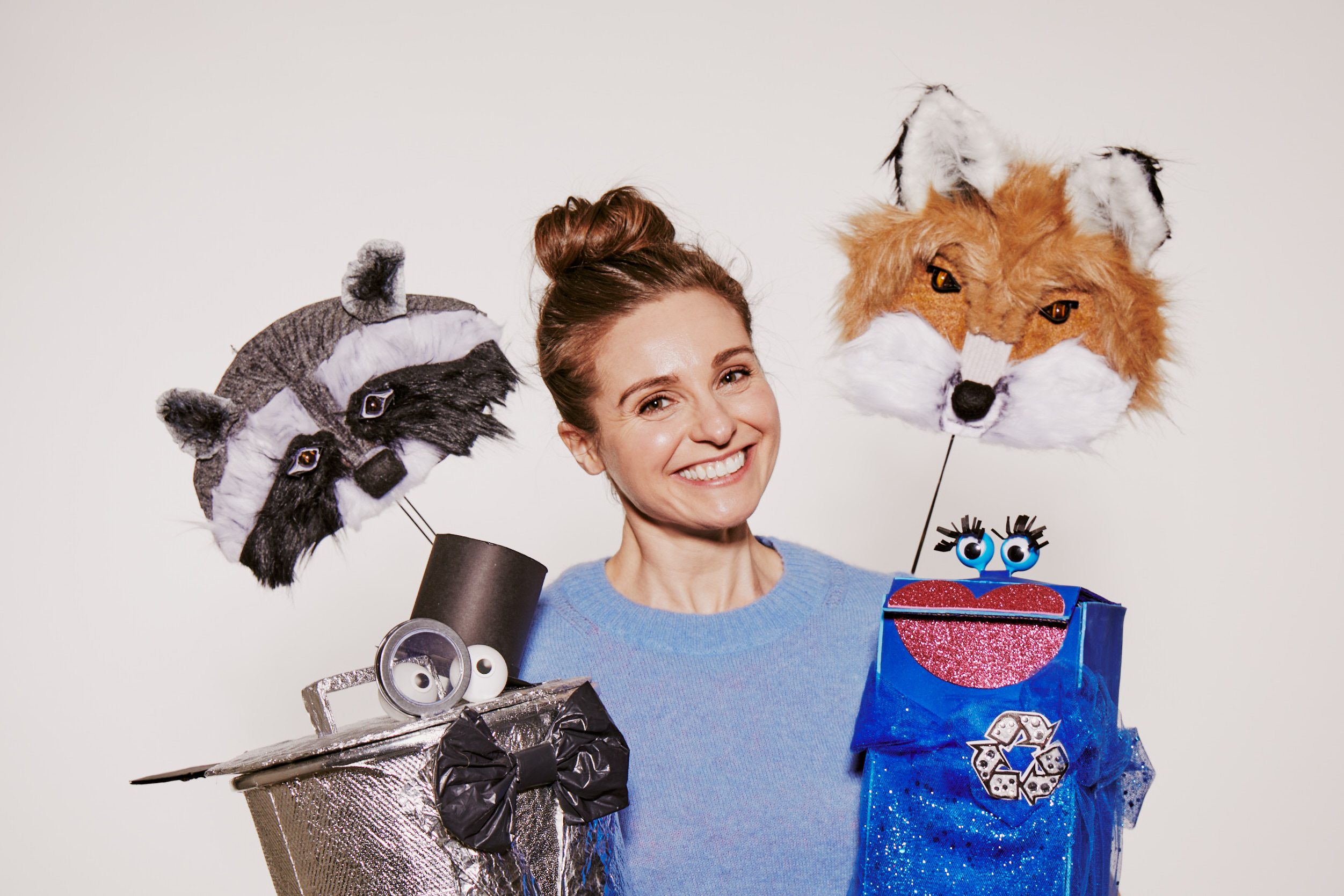 A person wearing a light blue sweater is holding two puppet heads on sticks. On the left is a raccoon puppet with a gray and black mask, and on the right is a fox puppet with orange and white fur. In front, a small robot puppet features a shiny metallic body with a top hat and bow tie, and a blue puppet with googly eyes and a sparkly heart. The background is plain, emphasizing the colorful and creative puppets.