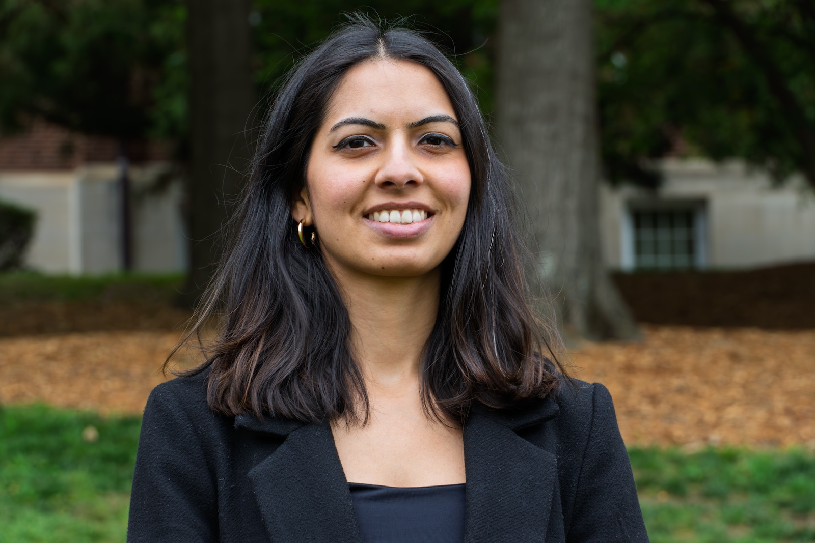 A woman looks at the camera smiling. There is a tree and grass behind her.