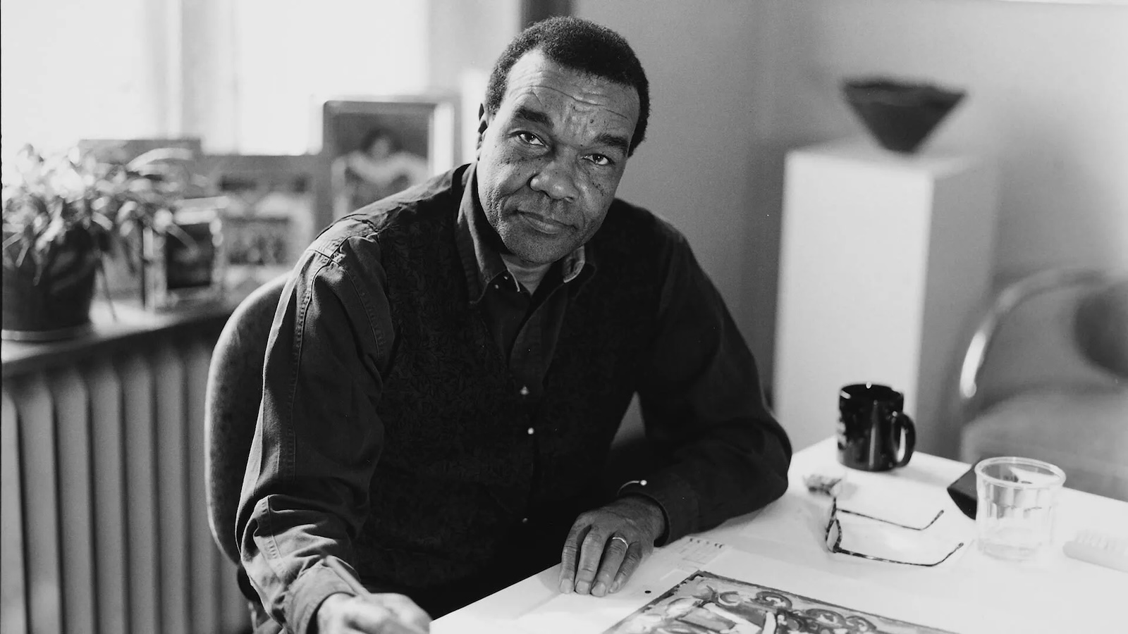 An artist looks directly at the camera. He holds a pen in his hand and a piece of art is in front of him on a table.