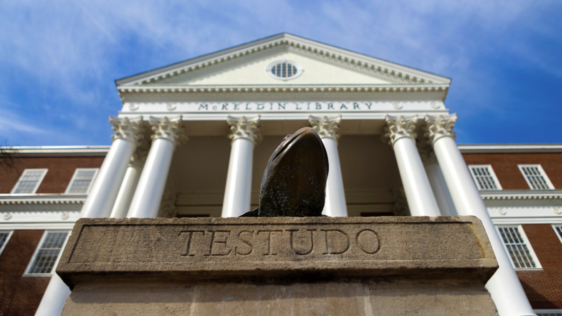 A photo of the testudo statue in front of McKeldin Library from a low perspective, capturing the name "TESTUDO" on the plinth, the columns of McKeldin Library, and a blue sky with light clouds.