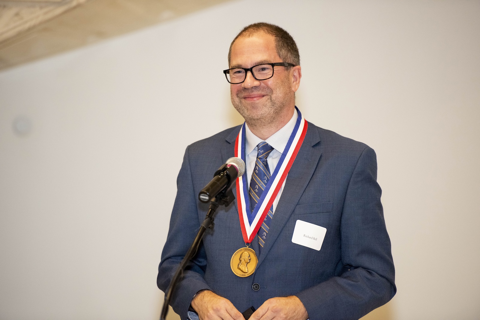 A man in a blue suit smiles. He wears an award around his neck.