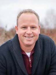 Headshot of a white man with short blonde and blue eyes hair smiling. He is wearing a blue sweater with a burgundy button down shirt underneath.