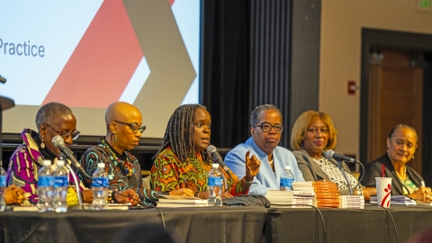 Dean Stephanie Shonekan sits on a panel of six Black women scholars, speaking into a microphone. The group is engaged in discussion during an academic event, with books and water bottles on the table in front of them.