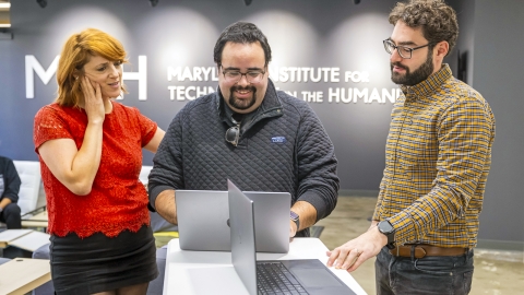 Three people gather around laptops at the Maryland Institute for Technology in the Humanities (MITH). They collaborate while smiling, with the MITH logo visible on the wall behind them.