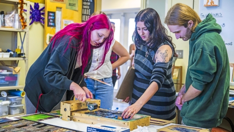 A student with bright pink hair demonstrates printmaking in a studio, rolling ink onto type while two other students watch closely. The workspace is lively, filled with posters and materials.