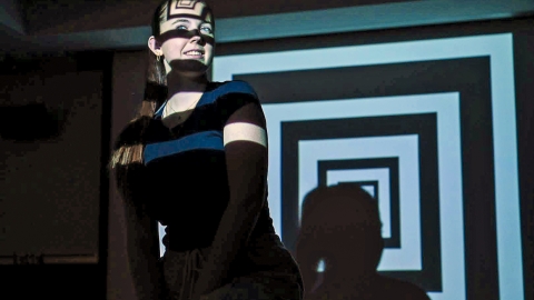 A student smiles while standing in front of a bold geometric light projection of concentric squares, the pattern casting shadows across her face and shirt.