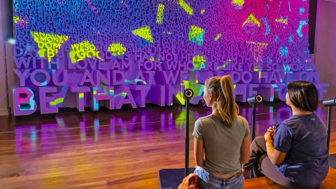 Two students sit on the floor in front of a large interactive art installation. Bright neon projections and bold 3D text fill the wall with words and phrases, creating an immersive digital environment.