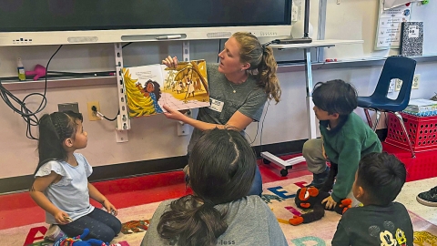 A woman sits on the floor of a classroom, holding open a colorful children’s book showing a girl reaching for bananas. Young children sit around her, engaged and attentive, symbolizing ARHU’s commitment to education and storytelling.