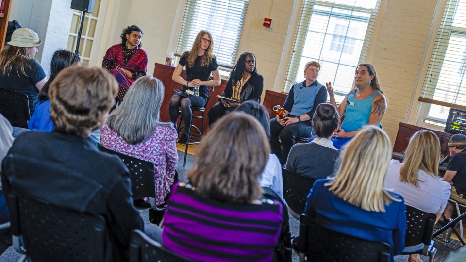 A panel of five speakers sits at the front of a bright room during a campus discussion. One participant in a turquoise top speaks while gesturing, as audience members seated in rows listen attentively.
