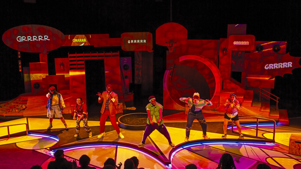 A cast of performers on stage act out a lively, colorful theater production under red and yellow lighting. Large speech bubbles reading “GRRRR” appear above them as they gesture dramatically in front of a set designed with oversized props.