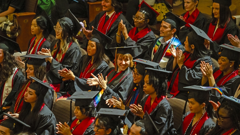 A large group of students in graduation caps and gowns sit in an auditorium, clapping and smiling during the commencement ceremony. Bright red stoles and honor cords add pops of color to the rows of graduates.