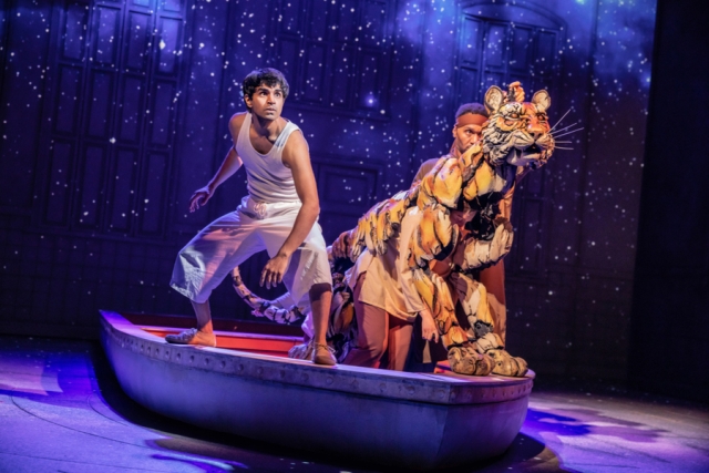 A theatrical scene featuring a performer in a white tank top and pants standing in a small boat alongside a large, lifelike tiger puppet.