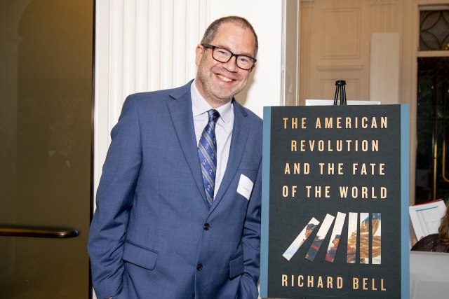A man in a suit smiles at the camera. He stands beside a book cover that says "The American Revolution and the Fate of the World"